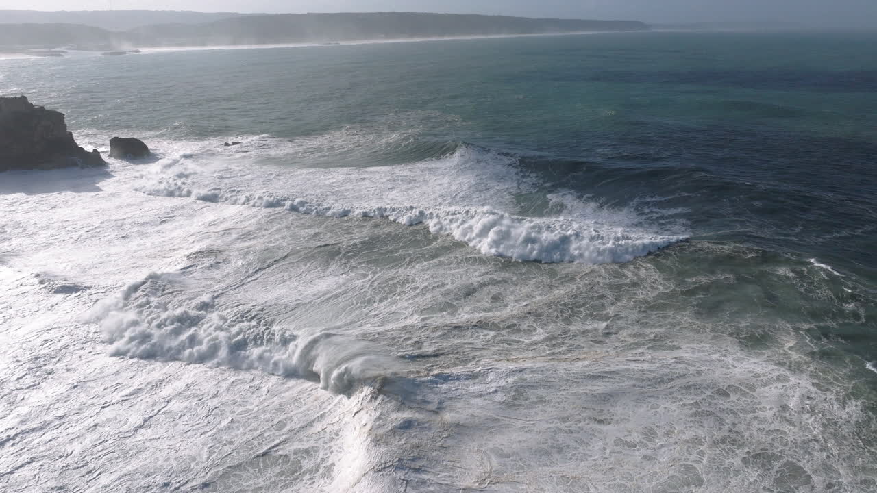 Aerial drone shot of waves coming into shore on a day with giant waves in Nazaré, Portugal, Europe. Farol da Nazaré lighthouse visible. Big wave surfing. Shot in 5K ProRes 422 HQ