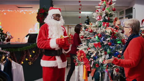 Trucking-shot-of-cheerful-man-impersonating-Santa-Claus-in-Christmas-adorn-shopping-mall-clothing-shop.-Supervisor-jingling-xmas-bells-and-chatting-with-elderly-client-in-holiday-themed-fashion-store