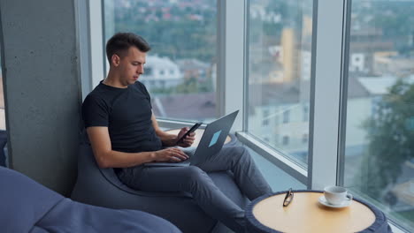 young man sitting in bean-bag chair with laptop on his knees. businessman looking at phone in his hands. cityscape at backdrop in blur.