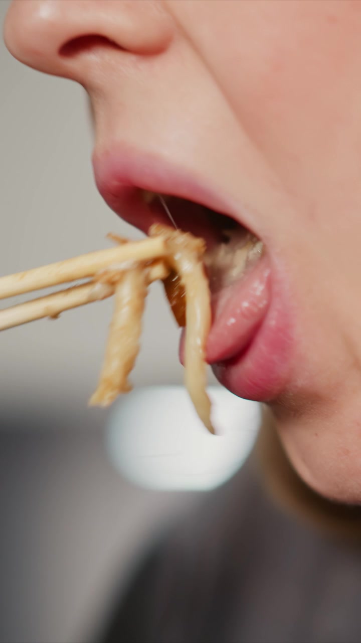 Close-up van een vrouw die noedels eet met eetstokjes, waarbij de focus ligt op haar mond en gezichtsuitdrukkingen. Het onderwerp brengt herhaaldelijk voedsel naar haar lippen met houten eetgerei tegen een onscherpe achtergrond.