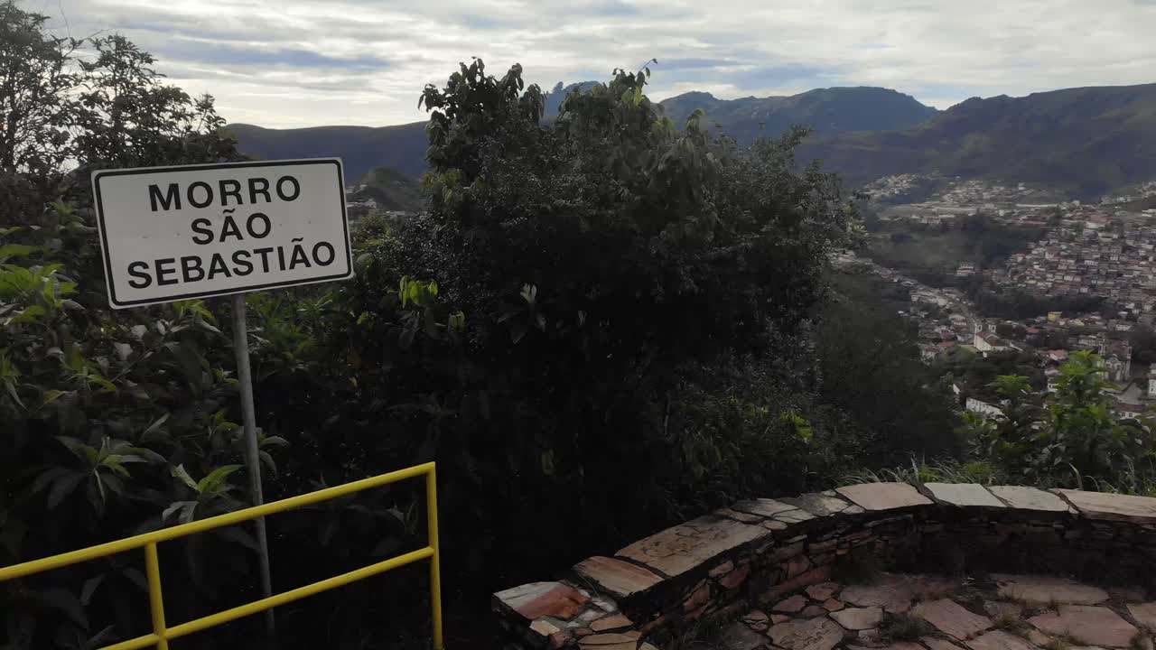 Sign of the Saint Sebastian hill viewpoint of Ouro Preto with Itacolomi mountain peak in the background slowly panning around seeing a bit of the historic city below. Translation: 'San Sebastian Hill'