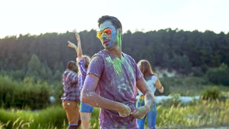 portrait of the young handsome man in sunglasses and all in colorful paints spots smiling to the camera and posing outdoors while having festive holi day with multiethnic friends