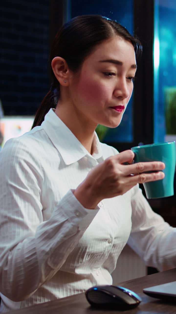 Video verticale. Un impiegato in ufficio beve un caffè mentre usa il laptop per fare ricerche per un progetto