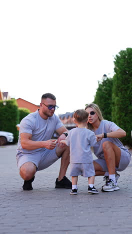 beautiful caucasian family outdoors. little baby stands at the parents sitting squatted near him. vertical video