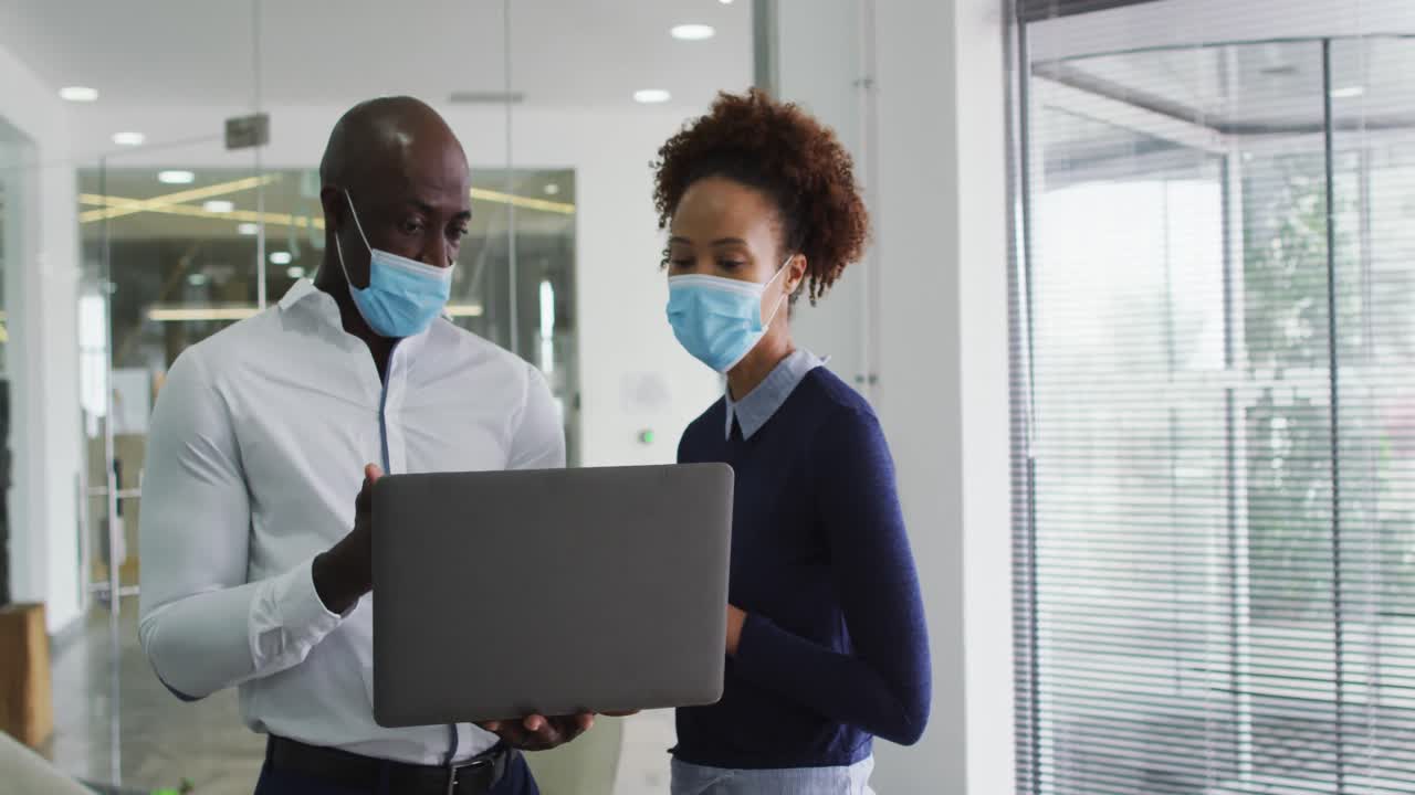 Diverse male and female business colleagues in face masks discussing, using laptop in office
