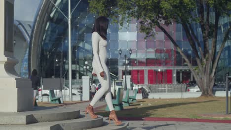 Una-Joven-Con-Un-Traje-Blanco-Disfruta-De-Un-Día-En-La-Ciudad-Con-Un-Edificio-De-Cristal-Detrás-De-Ella