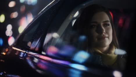stylish female is commuting home in a backseat of a taxi at night. beautiful woman passenger looking out of window while in a car in urban city street with working neon signs. cinematic footage.