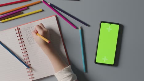 Fotografía-Cenital-De-Un-Niño-Con-Un-Teléfono-Móvil-De-Pantalla-Verde-Haciendo-La-Tarea-De-Matemáticas-En-La-Escuela-En-El-Libro-1