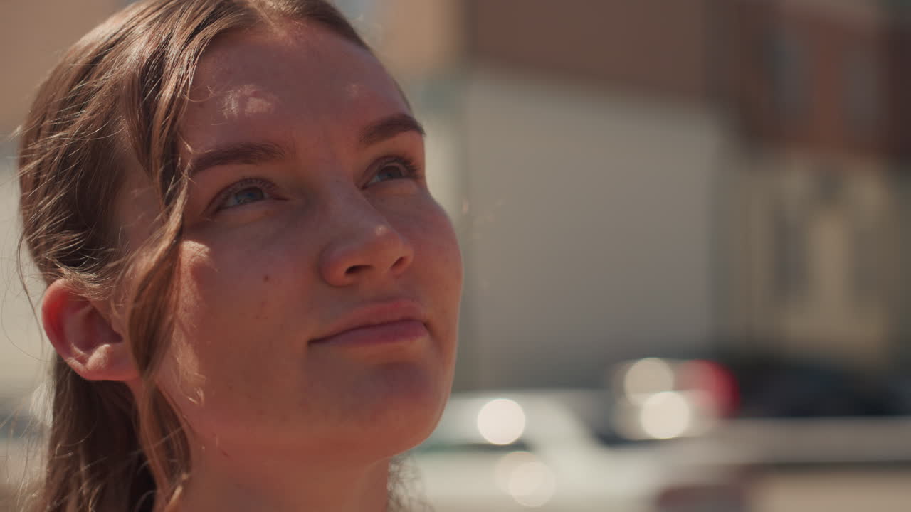 white woman in pink dress standing on sunlit city street, sunglasses on, soft smile, braided hair with loose curl, cars and brick buildings in background, warm golden light, candid portrait, hopeful