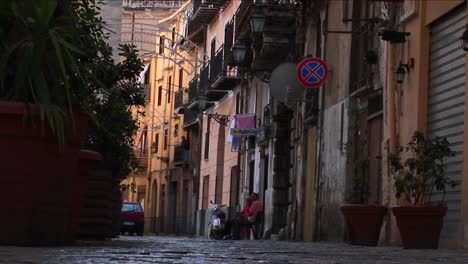 Un-Hombre-Se-Sienta-En-Una-Silla-Fuera-De-Los-Edificios-De-Apartamentos-Mientras-Los-Peatones-Caminan-Por-La-Calle-Palermo-Italia