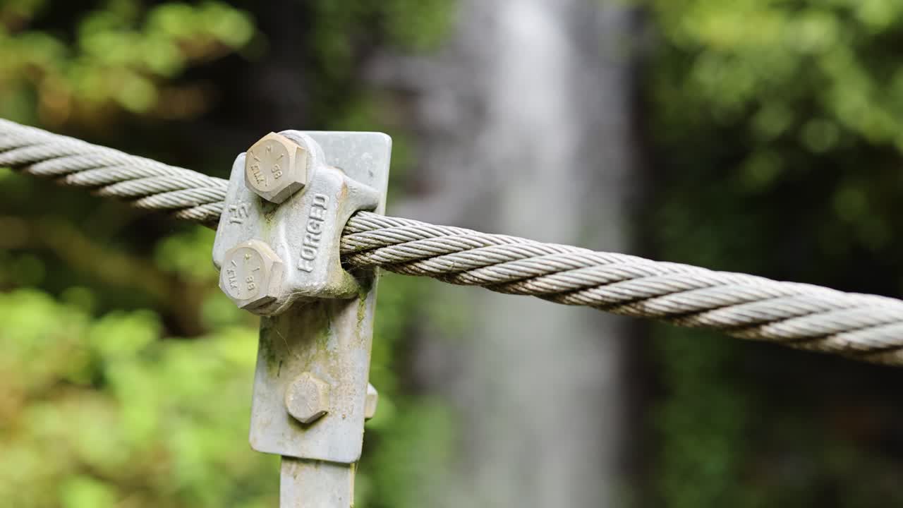 Close-up of a steel cable clamp against a blurred waterfall backdrop, highlighting industrial and natural contrast