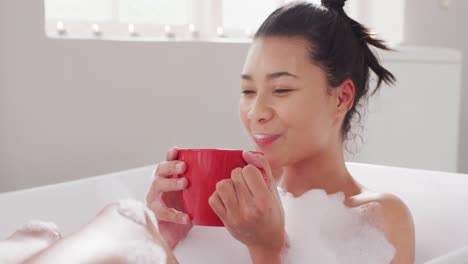 Video-of-portrait-of-smiling-biracial-woman-sitting-in-bathtub-in-bubble-bath-with-red-cup-of-tea