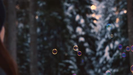 brunette-woman-with-winter-clothes-blowing-rainbow-colored-soap-bubbles-flying-suspended-in-the-air,-with-a-snowy-forest-in-the-background