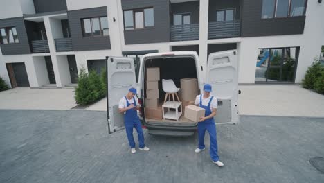 two young workers of removal company unload boxes and furniture from minibus into customer's home