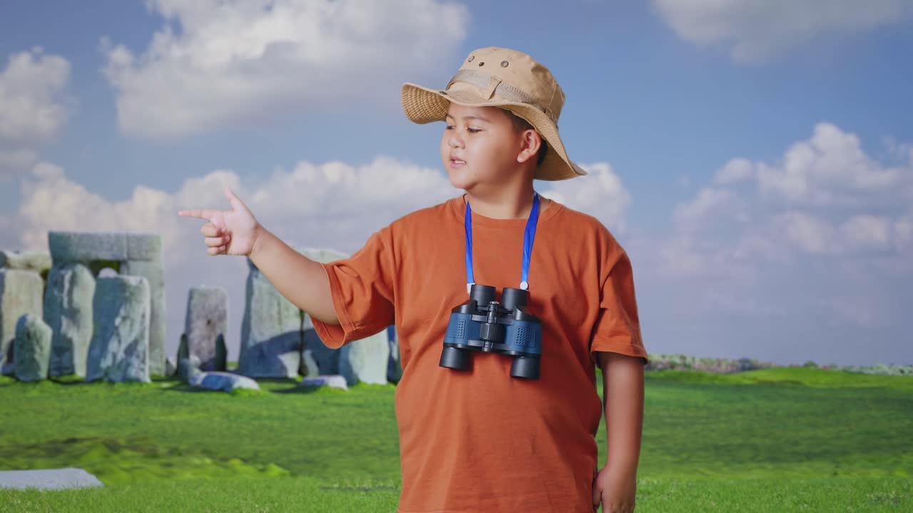 Asian Boy With A Hat And Binoculars Smiling And Pointing To Side While Traveling In Stonehenge. Boy Researcher Examines Something, Travel Tourism Adventure Concept