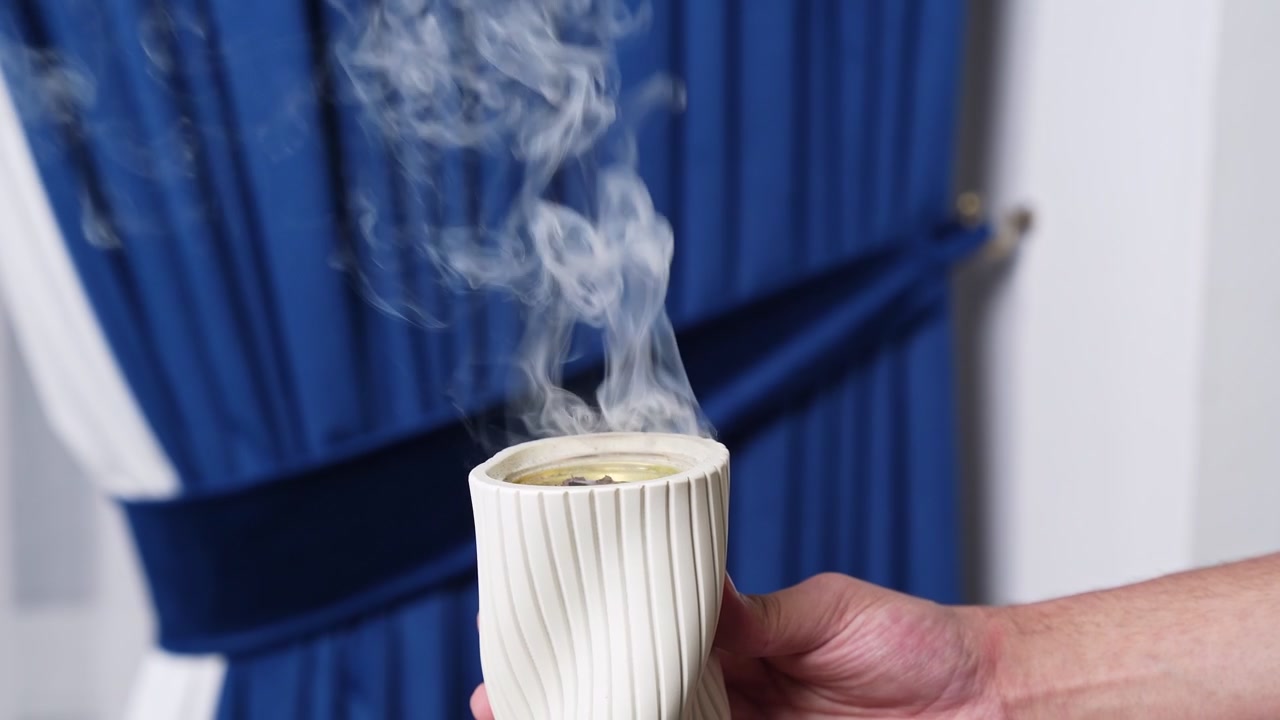 La mano de una persona sosteniendo un incensario de cerámica blanco retorcido con un denso humo aromático elevándose contra un fondo de cortina azul difuminada en un ambiente interior moderno