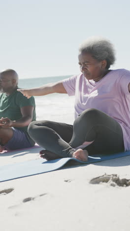 vertical video of senior african american couple doing yoga at beach, in slow motion