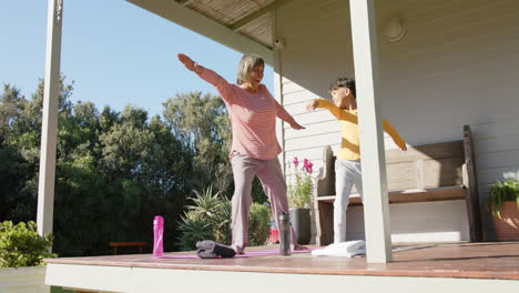 Senior-biracial-grandmother-and-grandson-doing-yoga-and-stretching-on-terrace-at-home,-slow-motion