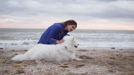 round camera movement: young woman sitting on the sand and embracing her dog of the samoyed breed by the sea. white fluffy pet