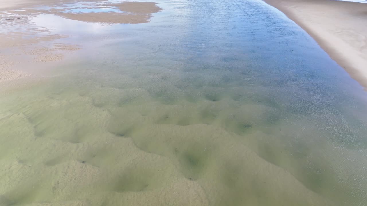 Drone glides above clear tidal channel, revealing sand ripples, shallow water, and bright daylight