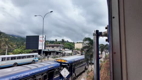 a view from a train in kandy, sri lanka, showcasing urban scenery with a road lined by buildings and lush greenery beneath a cloudy sky. captures travel and exploration essence.