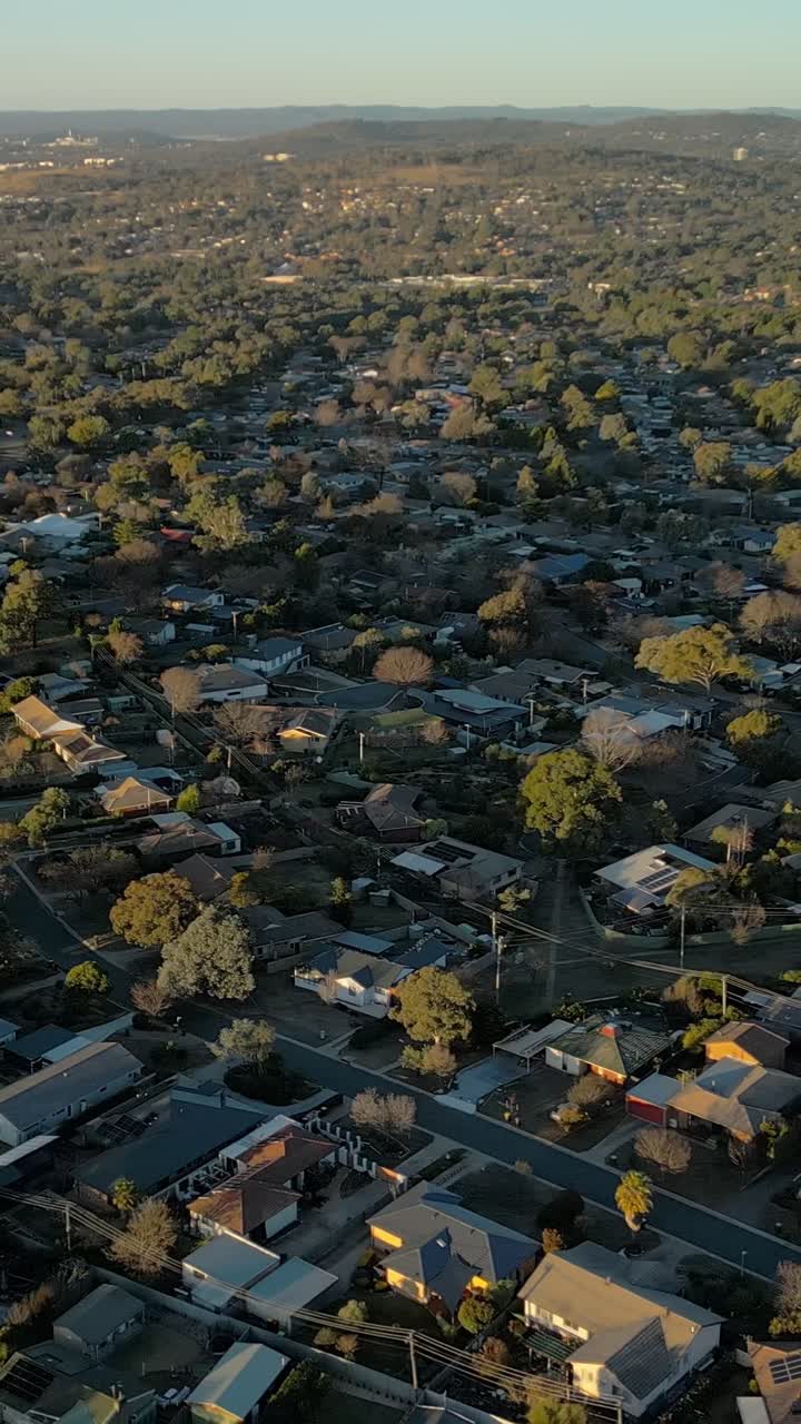 Un dron vertical vuela alrededor de calles suburbanas llenas de casas en el sur de Canberra, con la ciudad de Woden visible a la distancia. Zona residencial a última hora de la tarde mostrando el desarrollo urbano.