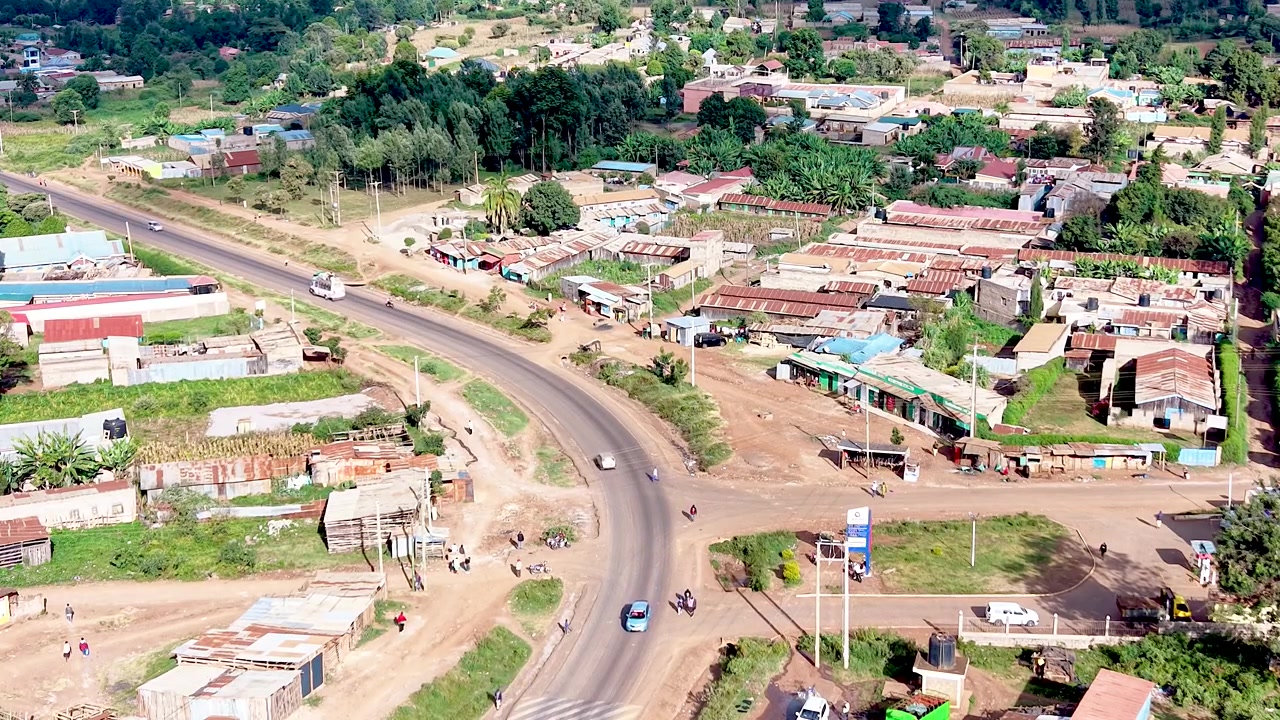 Una vista cinematografica del Kenya rurale che mostra insediamenti informali con tetti di lamiera arrugginiti, case logore, sentieri polverosi del villaggio e la vita quotidiana della comunità.