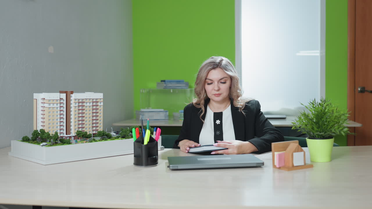 real estate professional seated in modern office holding notebook with thoughtful expression, surrounded by desk accessories, laptop, architectural model, and green indoor plant in bright workspace