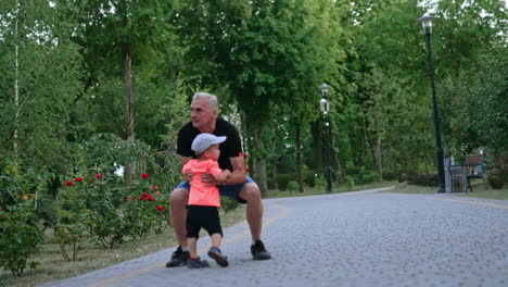 kid in cap runs up to his grandpa. man tosses the smiling baby boy and spins him in the park.