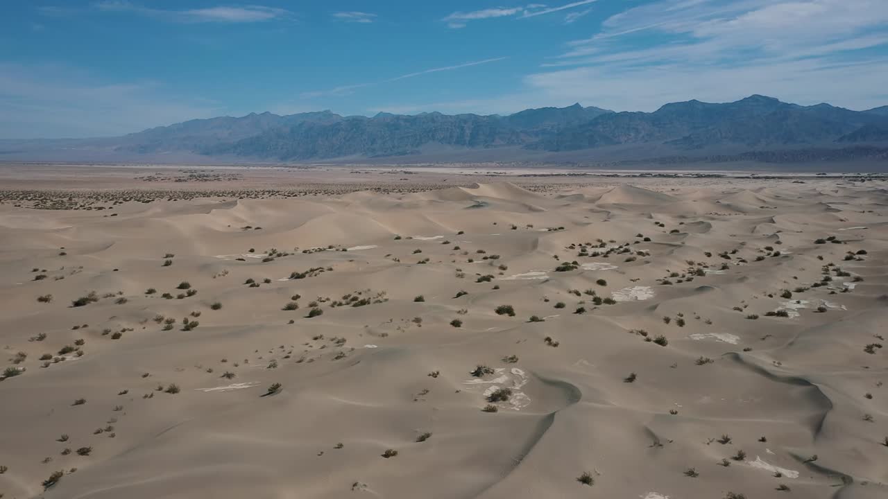 Aerial drone view showing vast sand dunes with scattered vegetation and distant mountain range under blue sky in Death Valley National Park, California, United States