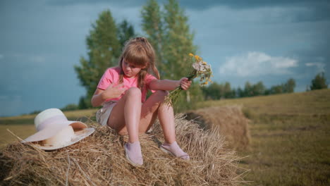 little girl sits on hay bale holding wildflowers while removing something from her arm, a white sun hat rests beside her as younger brother plays with hay bale in vast countryside