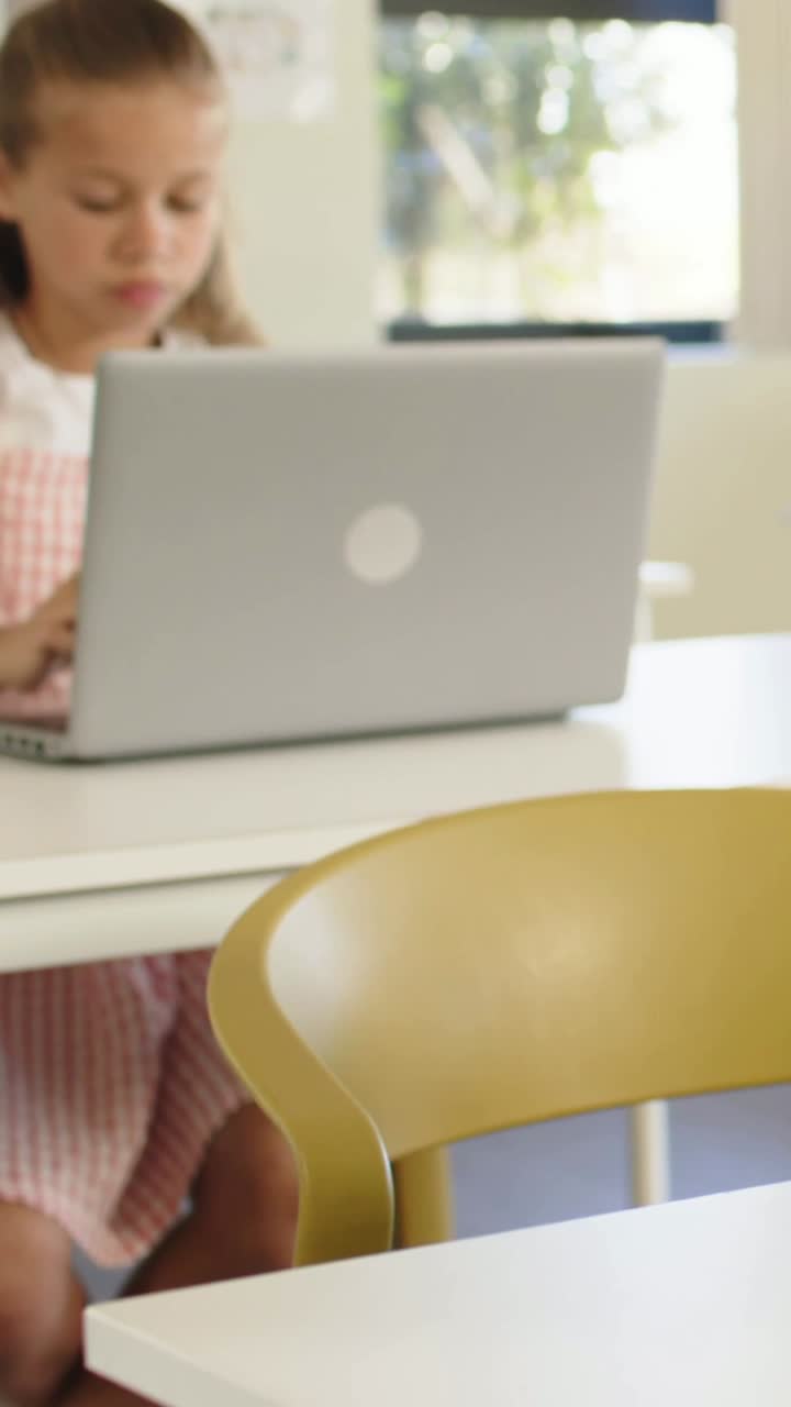 Vertical video: Opening child in pink typing for class at desk with laptop yellow chair, copy space