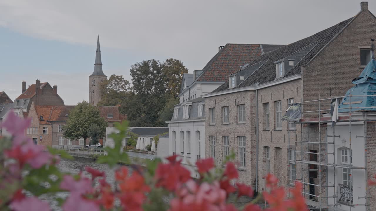 A picturesque, sunlit view along a historic canal in Bruges, Belgium, framed by traditional Flemish architecture with stepped-gables, leading the eye toward a distant, prominent city spire