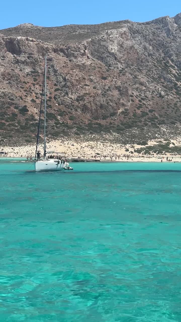 Yunanistan'da Ege Denizi kıyısında, kayalık dağ fonuyla kumlu plaj yakınında berrak turkuaz bir koyda demirlemiş yelkenlinin dikey manzarası.