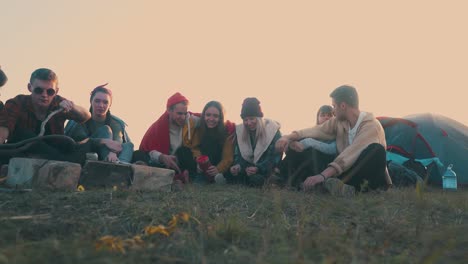 joyful tourists sit at burning bonfire on meadow at sunset