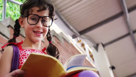 Smiling-schoolgirl-reading-book-in-campus-at-school