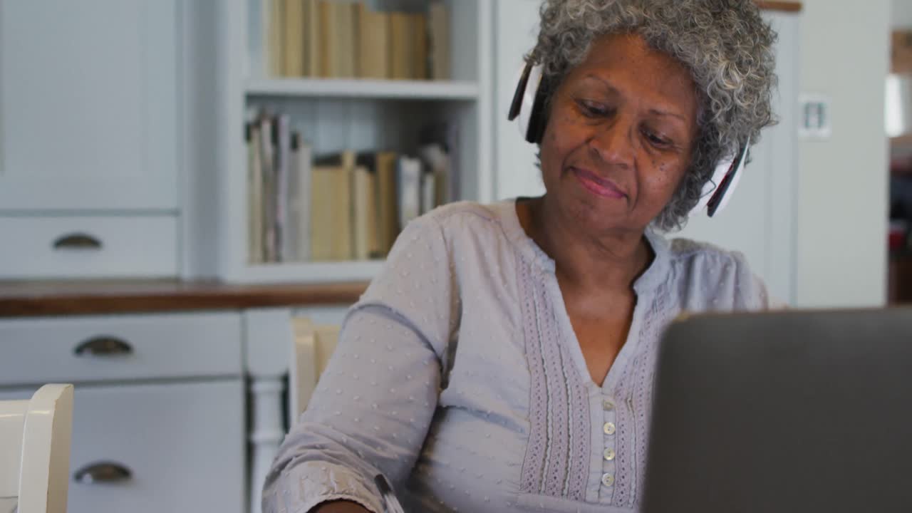 Senior african american woman wearing phone headset taking notes while using laptop at home