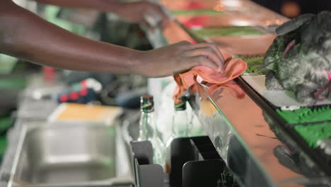 close-up of bartender's hand cleaning bar surface with orange cloth. plush rabbit sits on bar counter as green heineken neon lights glow in the background. bar tools and bottles are visible
