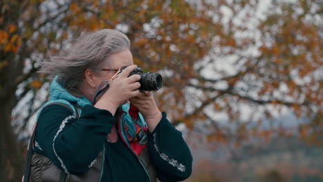 female photographer with grey hair and glasses taking pictures with her camera in nature, surrounded by orange leaved trees during a cold windy autumn day in slow motion