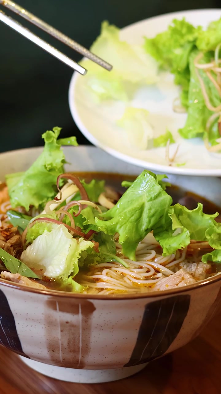 Primo piano di bacchette che aggiungono verdure fresche a una ciotola di ceramica di zuppa di noodle vietnamita.