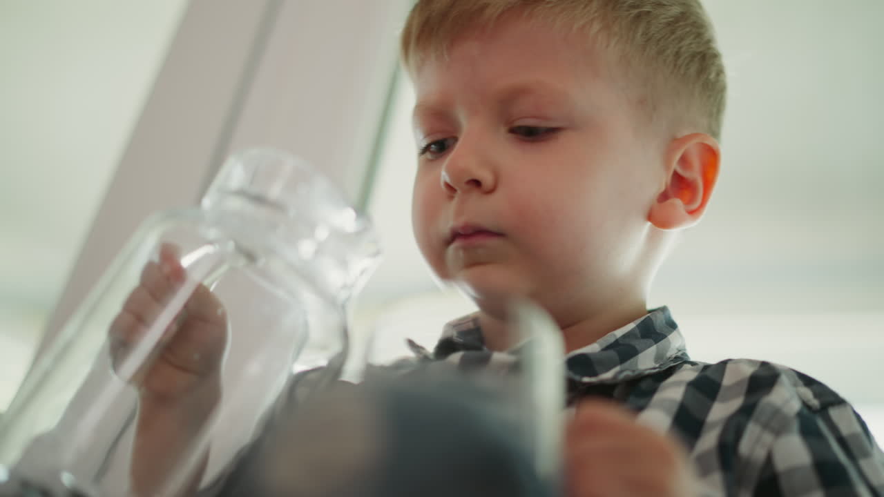close up of child sitting by window pouring water from transparent jar into glass cup placed on leg carefully concentrating on pouring process wearing checkered shirt