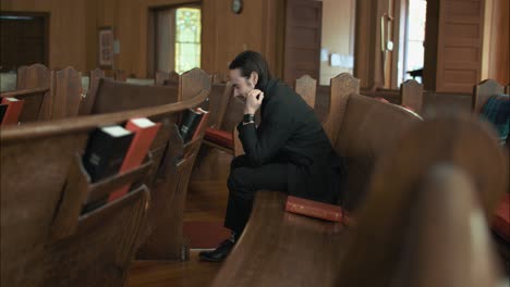 young man in black suit praying in church pew in cinematic slow motion with folded hands