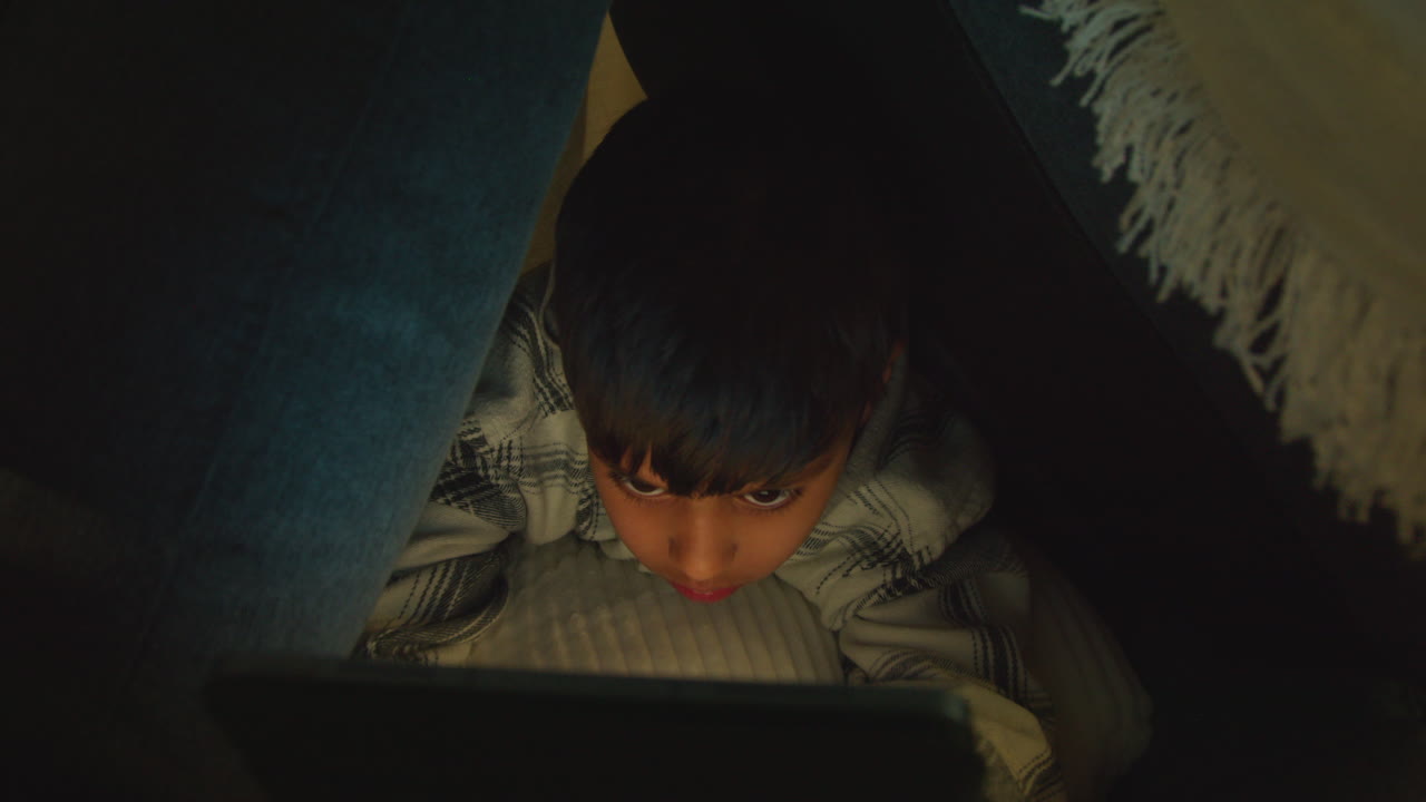 집에서 만든 어린 소년의 클로즈업, 밤에 디지털 태블릿으로 놀고 있는 베개로 만든 캠프 1