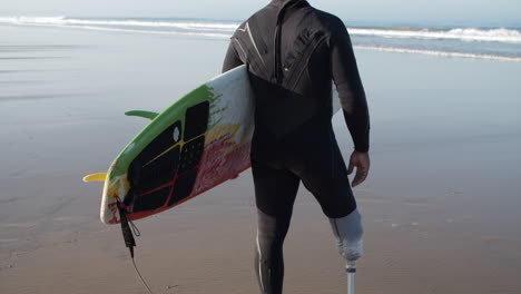 vista trasera de un surfista masculino con una pierna artificial parada en la orilla del océano y sosteniendo una tabla de surf bajo el brazo