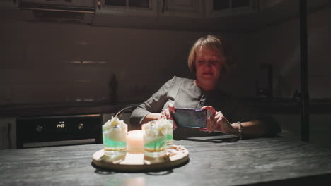 light focused on beautifully decorated cakes as baker leans in carefully snapping photos with her phone in dark cozy kitchen, capturing artistic dessert presentation illuminated by soft candlelight