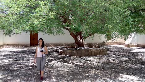 una niña caminando bajo un gran árbol en un patio histórico, sosteniendo su cámara