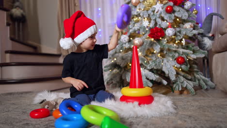beautiful baby boy in red cap playing with pyramid. kid plays with toy near the beautiful christmas tree.