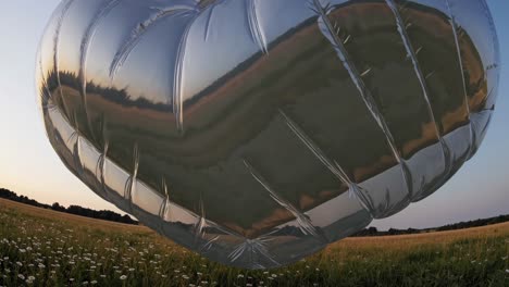 gran globo de corazón de mylar plateado flotando sobre un campo de flores blancas durante una hermosa puesta de sol, reflejando el cielo colorido y el paisaje