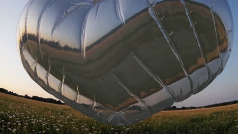 large silver mylar heart balloon floating over a field of white flowers during a beautiful sunset, reflecting the colorful sky and landscape