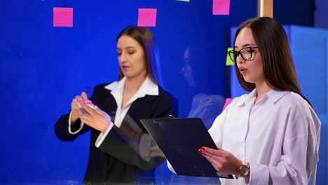 two ladies standing at the glass wall talking and putting stickers on it. co-workers communicate pointing at sticky notes.
